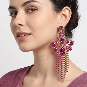 A woman wearing a large ornate Fuchsia crystal statement earring on gold with cascading bead fringe and clear crystal accents against a light gray background.