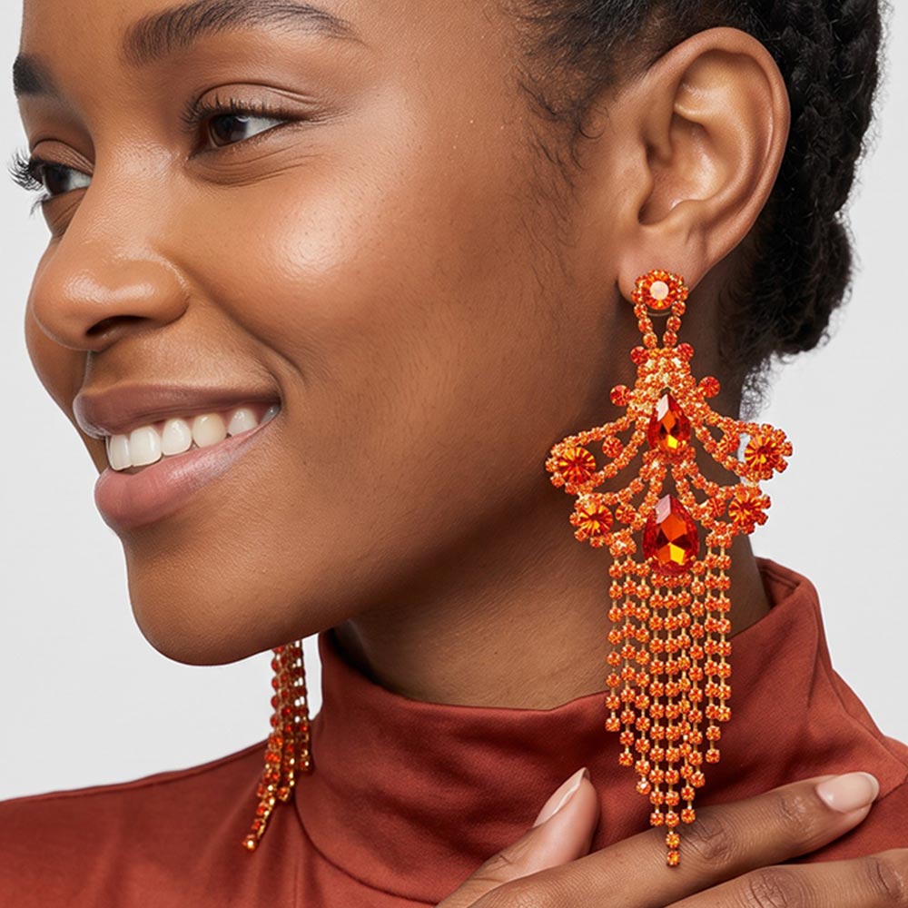 A woman wearing a large ornate Orange crystal statement earring on gold with cascading bead fringe and clear crystal accents against a light gray background.