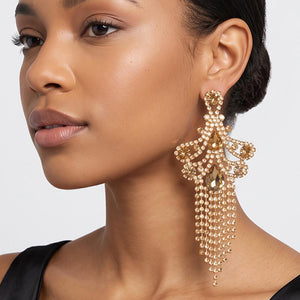 A woman wearing a large ornate Champagne Gold Stone statement earring with cascading bead fringe and clear crystal accents against a light gray background.