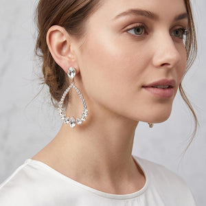 A close-up of a woman wearing a large silver teardrop dangle earring adorned with clear crystals, set against a soft light gray background.