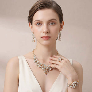 A woman models a matching 4-piece jewelry set on gold with statement necklace, ring, and bracelet adorned with champagne gold stone crystals, paired with matching drop earrings. She is wearing a cream-colored sleeveless dress against a soft beige background.