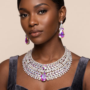 AB iridescent crystal statement necklace set on silver-tone metal, featuring a broad, layered bib of marquise and round stones with a large teardrop centerpiece and matching teardrop earrings reflecting rainbow colors. Necklace measures 17 inches plus a 2-inch extender, with 2.25-inch decor; earrings are 2.2 inches long. Worn by a woman in a velvet charcoal outfit with wide shoulder straps against a warm beige background.
