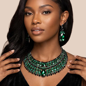 Emerald green crystal statement necklace set on silver-tone metal, featuring a broad, layered bib of marquise and round stones with a large teardrop centerpiece and matching teardrop earrings. Necklace measures 17 inches plus a 2-inch extender, with 2.25-inch decor; earrings are 2.2 inches long. Worn by a woman in a black V-neck outfit against a warm beige background.