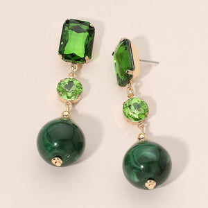 Drop earring with a green rectangular stone, a smaller light green round stone, and a larger dark green teardrop stone, set in gold-toned metal against a soft pink background.