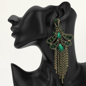 A large ornate Emerald Green crystal statement earring on gold with cascading bead fringe and clear crystal accents on a black earring mannequin.