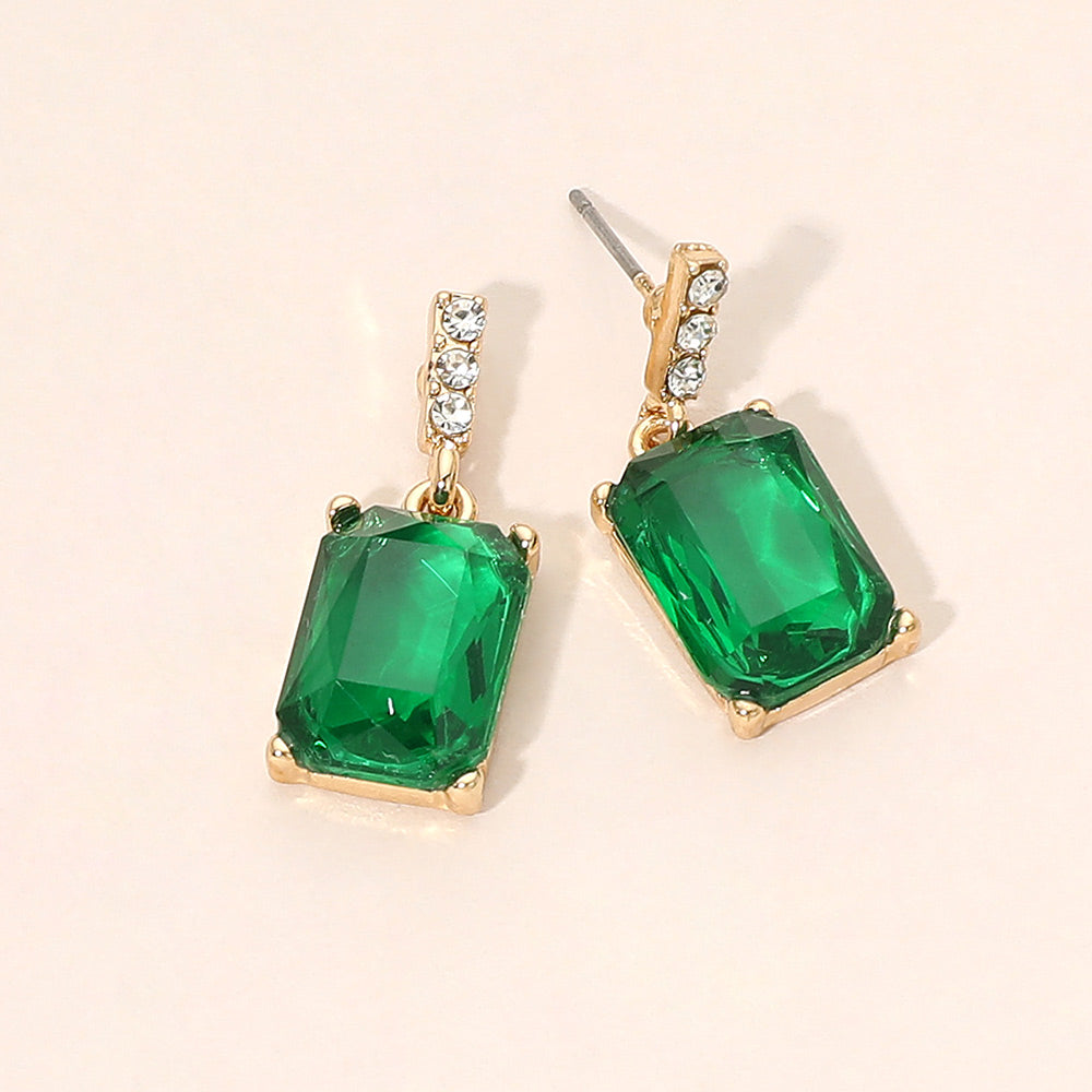 A pair of gold-tone drop earrings featuring large rectangular emerald green crystals with a short row of small clear crystals above each stone, displayed on a light beige background.