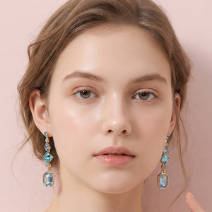 A close-up of a model wearing blue geometric crystal link drop earrings on gold with faceted stones on a beige background.