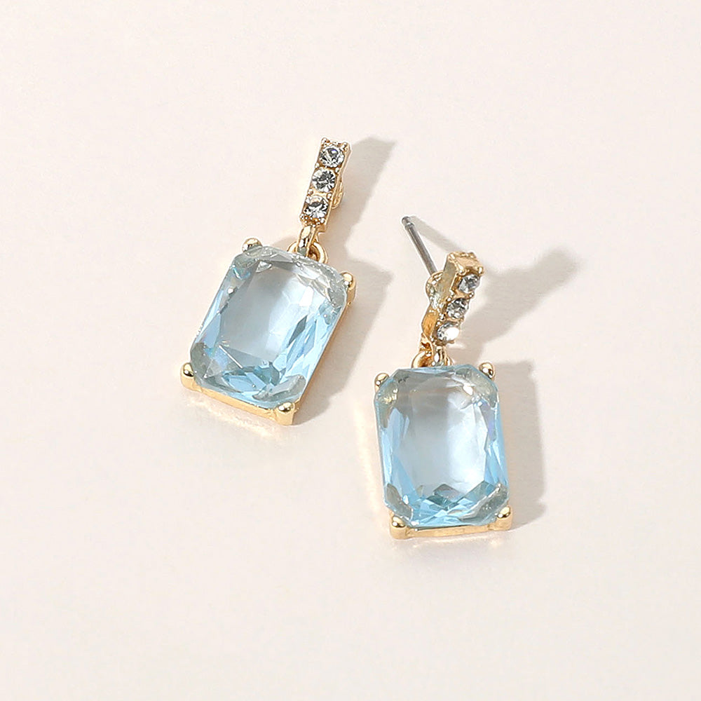 Gold-tone dangle earrings featuring large rectangular light blue crystals with a short row of small clear crystals above each stone, displayed on a light beige background.
