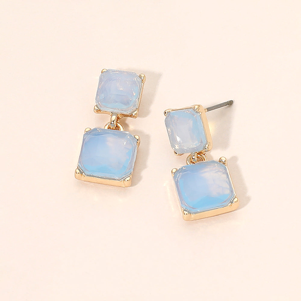 Blue opal double-square drop earrings on gold-tone metal, each featuring a smaller square stone connected to a larger square stone below. Each earring measures approximately 0.4 inches wide by 0.9 inches long. Displayed on a pale peach surface.