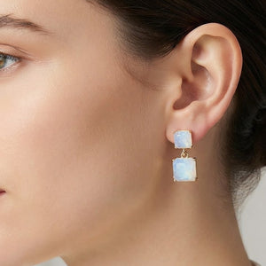 Blue opal double-square drop earring featuring a smaller pale blue square stone above a larger square drop, measuring approximately 0.4 inches wide by 0.9 inches long. Shown on a woman’s ear with her hair pulled back against a softly blurred neutral background to illustrate the style’s scale.
