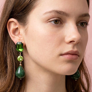 A close-up of a woman wearing a drop earring with a green rectangular stone, a smaller light green round stone, and a larger dark green teardrop stone, set in gold-toned metal against a soft pink background.