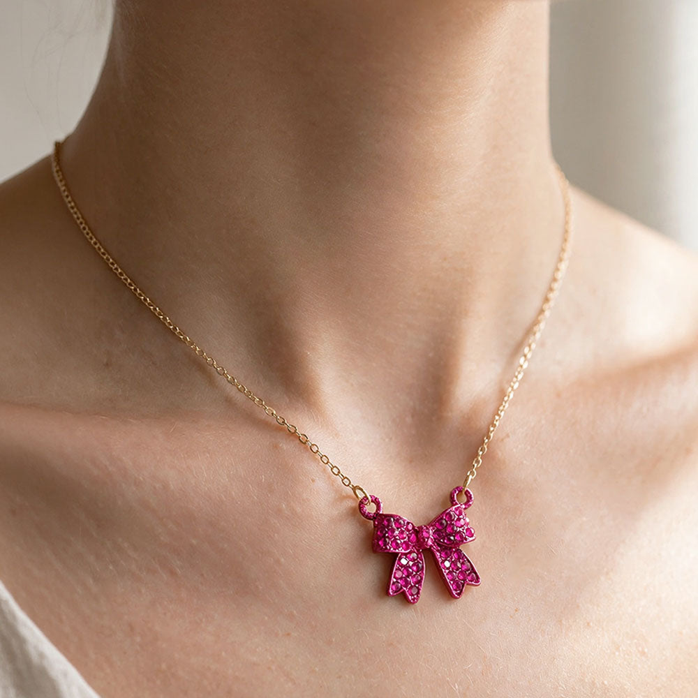 Bow pendant necklace with fuchsia hot pink crystals in a matching pink setting on a gold-tone chain, measuring 15.5 inches with a 2-inch extender. The bow measures 0.75 inch wide by 0.75 inch long, with two rounded loops, a center knot, and two ribbon tails covered with small round crystals, displayed on a white jewelry card.