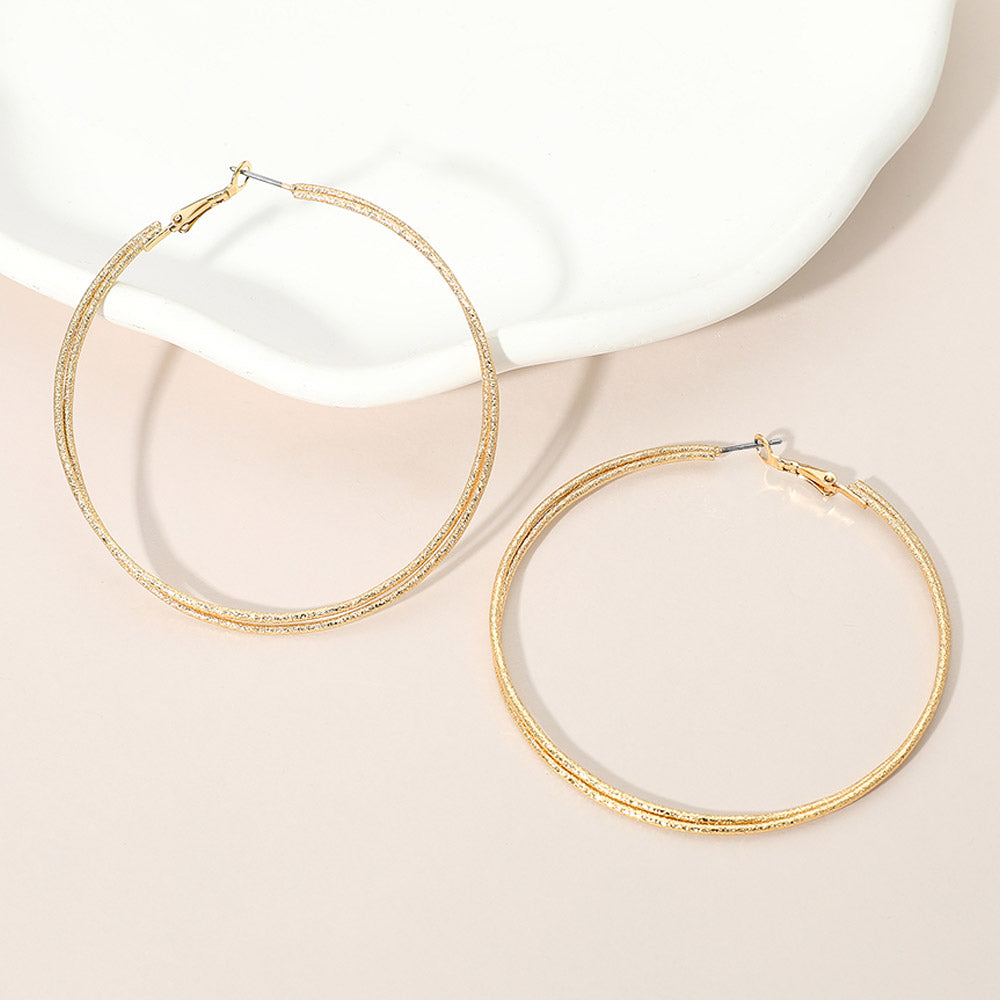 Gold textured double hoop earrings with a 2.75-inch diameter and pin catch clasp, displayed on a light pink background.