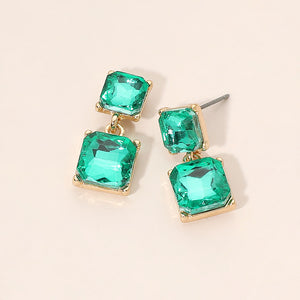 Green crystal double-square drop earrings on gold-tone metal, each featuring a smaller faceted square stone connected to a larger square stone below. Each earring measures approximately 0.4 inches wide by 0.9 inches long. Displayed on a pale peach surface.