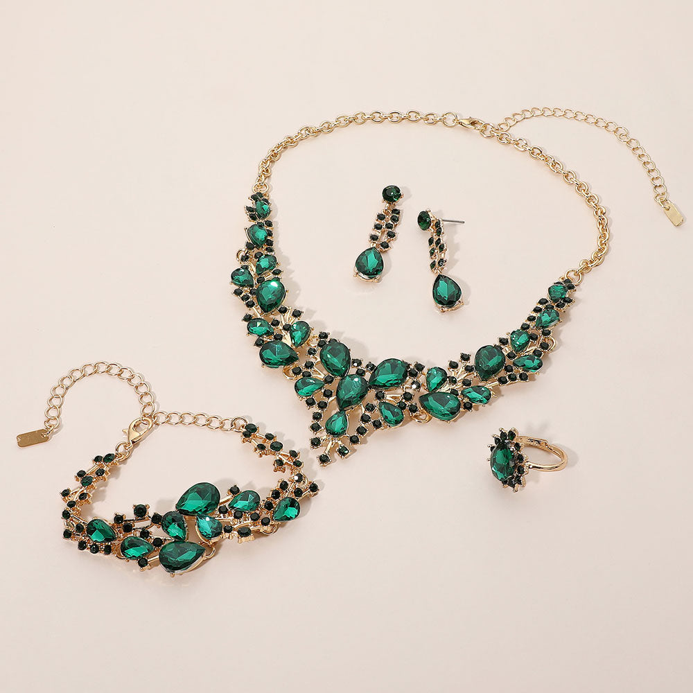 Emerald green crystal 4-piece jewelry set on gold with statement necklace, matching bracelet, drop earrings and adjustable ring arranged on a light beige background.