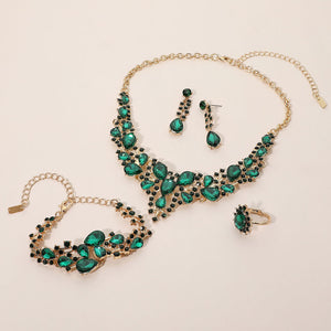 Emerald green crystal 4-piece jewelry set on gold with statement necklace, matching bracelet, drop earrings and adjustable ring arranged on a light beige background.
