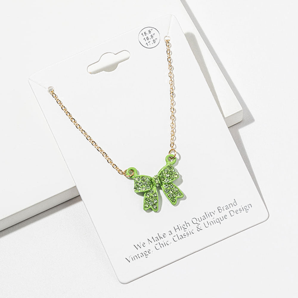Bow pendant necklace with green crystals in a green setting on a gold-tone chain, measuring 15.5 inches with a 2-inch extender. The bow measures 0.75 inch wide by 0.75 inch long, with two rounded loops, a center knot, and two ribbon tails covered with small round crystals, displayed on a white jewelry card.