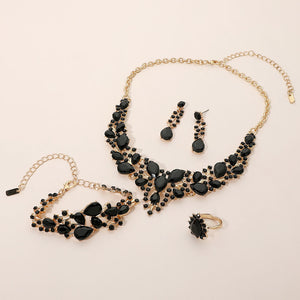 Jet black crystal 4-piece jewelry set on gold with statement necklace, matching bracelet, drop earrings and adjustable ring arranged on a light beige background.