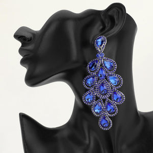 An extra-long ornate drop earring with cascading sapphire royal blue teardrop-shaped crystals and silver-tone metal detailing, displayed on a black mannequin head against a light gray background.