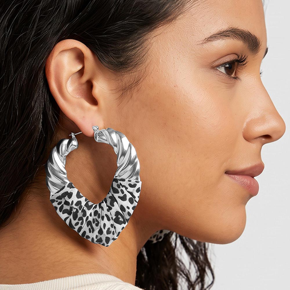 A close-up side profile of a woman wearing large twisted hoop earrings with a leopard print pattern and silver-tone metal edges on a light gray background.