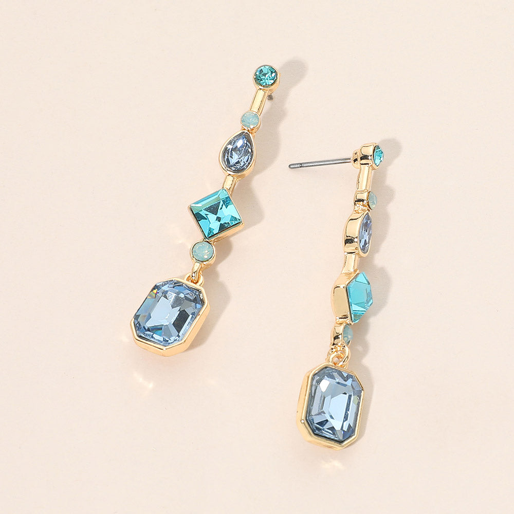 A pair of gold-plated geometric dangle earrings with light blue faceted gemstones in various shapes, displayed on a warm beige background.