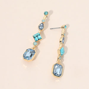 A pair of gold-plated geometric dangle earrings with light blue faceted gemstones in various shapes, displayed on a warm beige background.