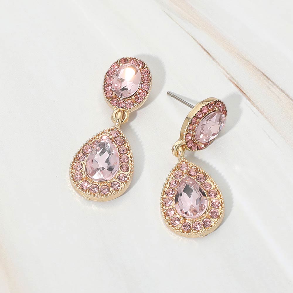 A pair of gold-tone teardrop dangle earrings with light rose pink crystal center stones surrounded by smaller pink rhinestones, displayed on a white marble surface.