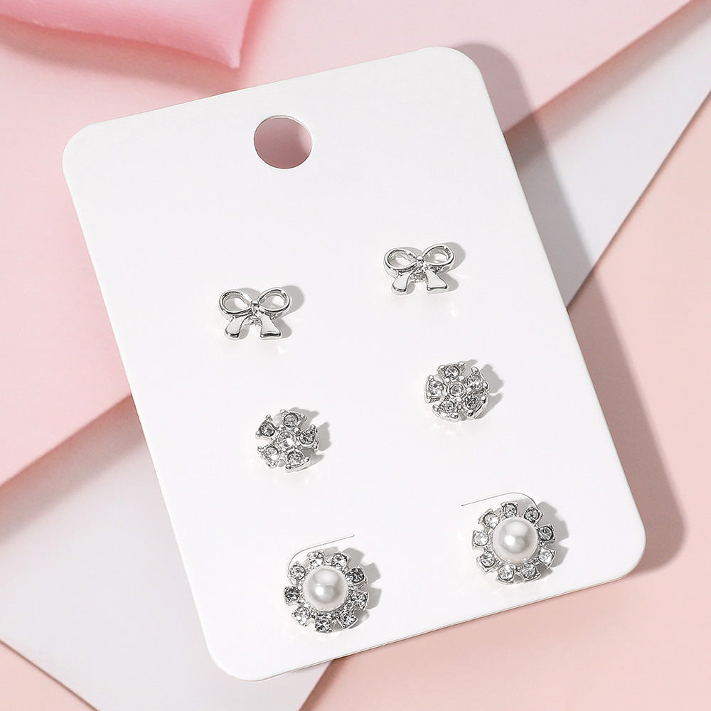 Little girls three-pair stud earring set in a silver finish featuring bow studs, round crystal cluster studs, and pearl halo studs, displayed on a white jewelry card with a pink background.
