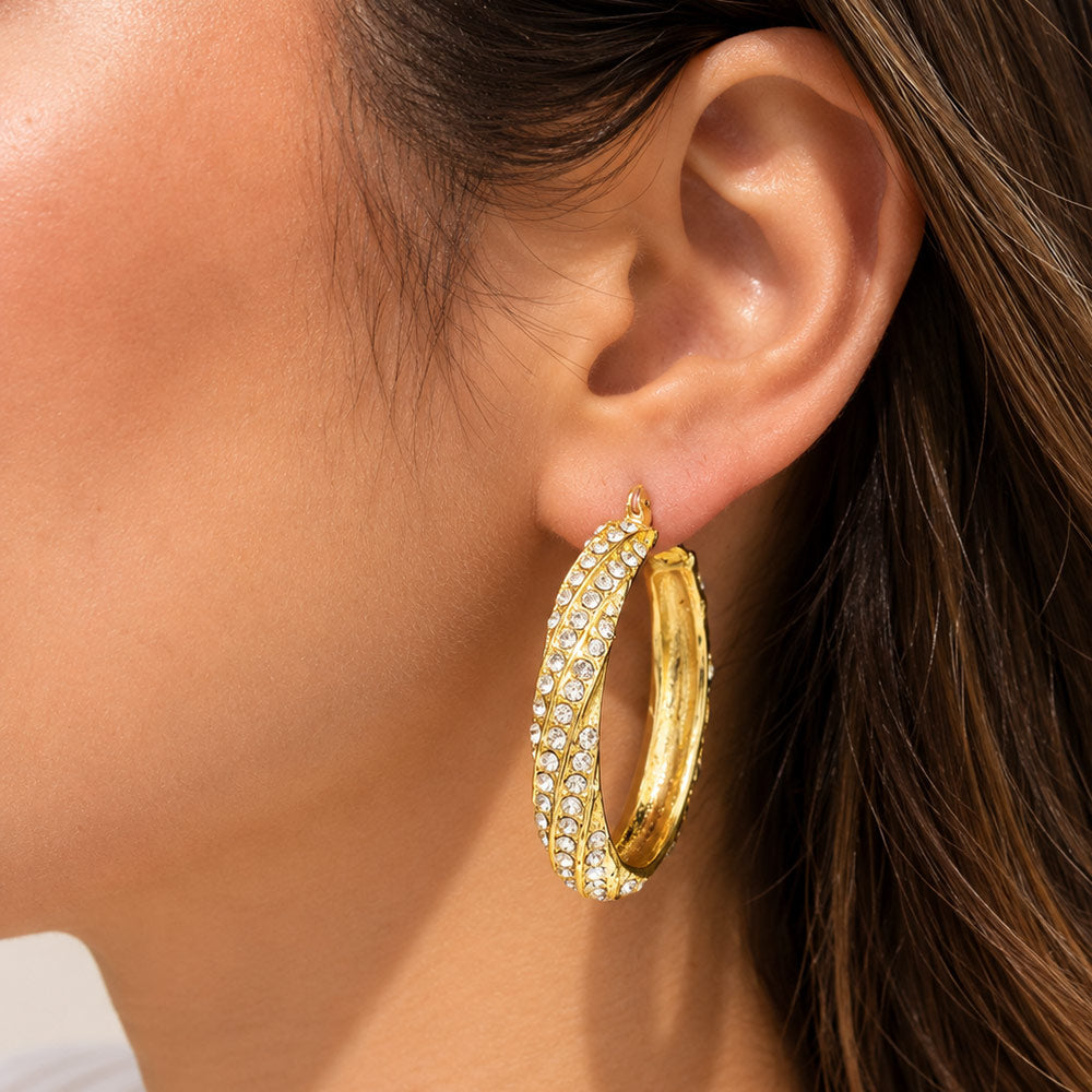 A close-up of a model's ear wearing a pair of gold hoop earrings adorned with rows of clear pavé stones.