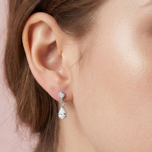 A close-up of a model's ear wearing a silver drop earring with a small round clear stone and a teardrop-shaped clear stone below, silver backed.