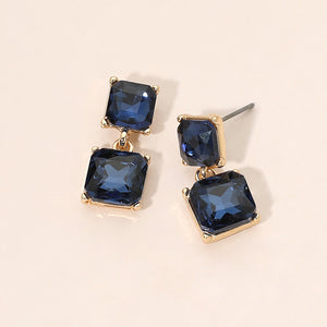 Navy crystal double-square drop earrings on gold-tone metal, each featuring a smaller faceted square stone connected to a larger square stone below. Each earring measures approximately 0.4 inches wide by 0.9 inches long. Displayed on a pale peach surface.