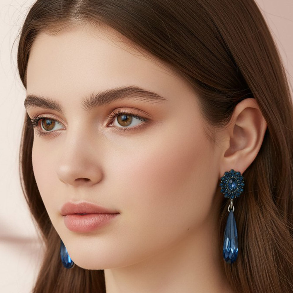 A close-up of a woman wearing a pair of navy teardrop dangle earrings with textured circular floral tops, backed in gun metal, shown against a soft beige background.