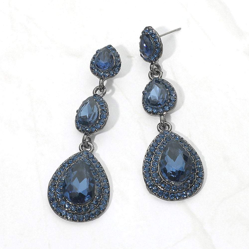 A pair of gunmetal drop earrings with three cascading teardrop-shaped navy gemstones, each surrounded by a halo of small dark rhinestones, displayed on a light textured background.