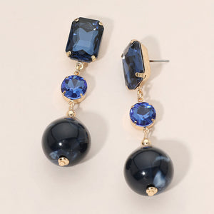 Navy faceted crystal and ball drop statement earrings on gold with rectangular, round, and spherical navy stones, displayed on a light beige background.