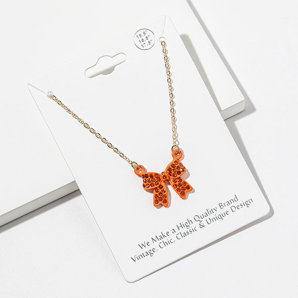 Bow pendant necklace with orange crystals in an orange setting on a gold-tone chain, measuring 15.5 inches with a 2-inch extender. The bow measures 0.75 inch wide by 0.75 inch long, with two rounded loops, a center knot, and two ribbon tails covered with small round crystals, displayed on a white jewelry card.