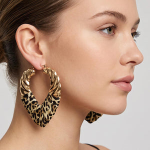 A close-up side profile of a woman wearing large twisted hoop earrings with a leopard print pattern and gold-tone metal edges on a light gray background.