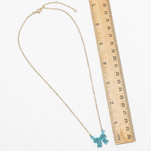 Bow pendant necklace with aqua crystals on a gold-tone chain, arranged in a long loop beside a wooden ruler. Necklace measures 15.5 inches around with a 2-inch extender; the bow measures 0.75 inch wide and long, displayed on a white surface.