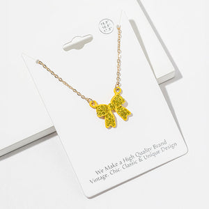 Bow pendant necklace with yellow crystals in a yellow setting on a gold-tone chain, measuring 15.5 inches with a 2-inch extender. The bow measures 0.75 inch wide by 0.75 inch long, with two rounded loops, a center knot, and two ribbon tails covered with small round crystals, displayed on a white jewelry card.