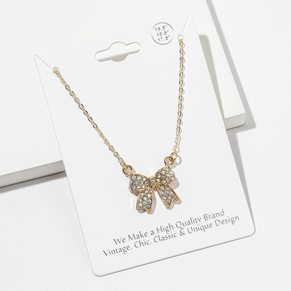 Bow pendant necklace with iridescent Aurora Borealis crystals on a gold-tone setting and chain, measuring 15.5 inches with a 2-inch extender. The bow measures 0.75 inch wide by 0.75 inch long, with two rounded loops, a center knot, and two ribbon tails covered with small round crystals, displayed on a white jewelry card.