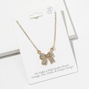 Bow pendant necklace with iridescent Aurora Borealis crystals on a gold-tone setting and chain, measuring 15.5 inches with a 2-inch extender. The bow measures 0.75 inch wide by 0.75 inch long, with two rounded loops, a center knot, and two ribbon tails covered with small round crystals, displayed on a white jewelry card.