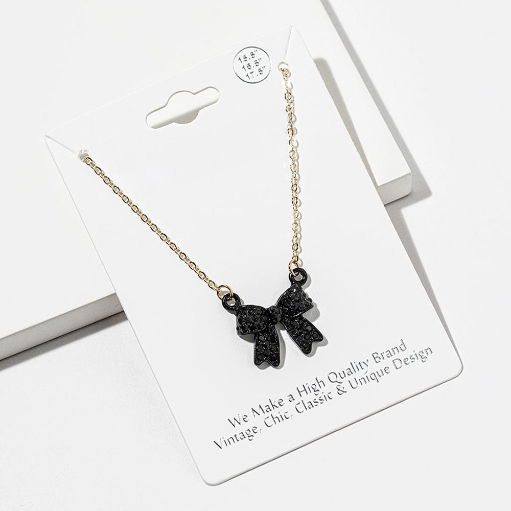 Bow pendant necklace with black crystals in a black setting on a black chain, measuring 15.5 inches with a 2-inch extender. The bow measures 0.75 inch wide by 0.75 inch long, with two rounded loops, a center knot, and two ribbon tails covered with small round crystals, displayed on a white jewelry card.