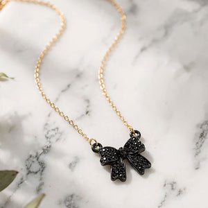 Close up of Bow pendant necklace with black crystals in a black setting on a black chain, measuring 15.5 inches with a 2-inch extender. The bow measures 0.75 inch wide by 0.75 inch long, with two rounded loops, a center knot, and two ribbon tails covered with small round crystals, displayed on white marble.