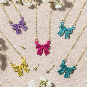 Five crystal bow pendant necklaces on gold-tone chains in purple, bright pink, yellow, light blue, and teal. Each 0.75-inch bow has crystal-covered loops, a center knot, and two ribbon tails, arranged on beige fabric among small white flowers.