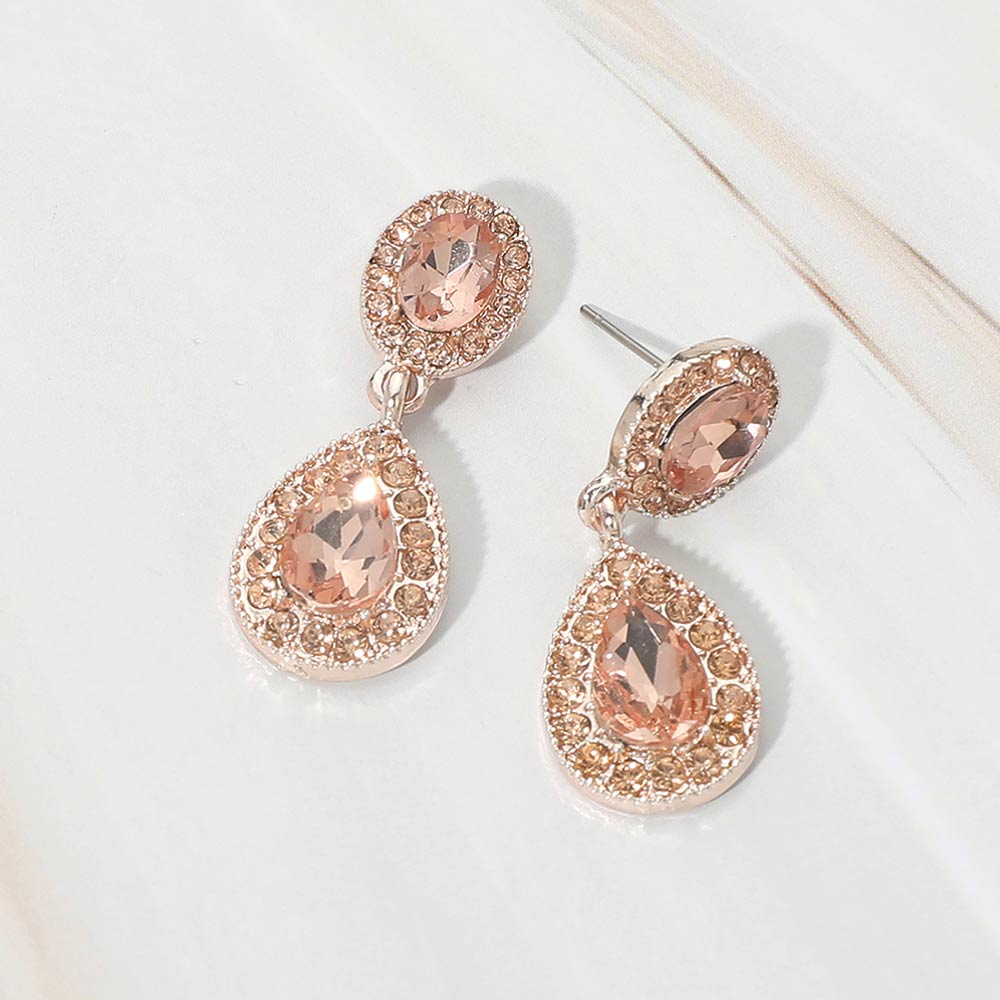 A pair of rose gold-tone dangle earrings with oval peach crystal top stones and teardrop peach crystal drops framed by small sparkling rhinestones, displayed on a marble surface.