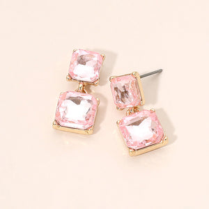 Pink crystal double-square drop earrings on gold-tone metal, each featuring a smaller faceted square stone connected to a larger square stone below. Each earring measures approximately 0.4 inches wide by 0.9 inches long. Displayed on a pale peach surface.