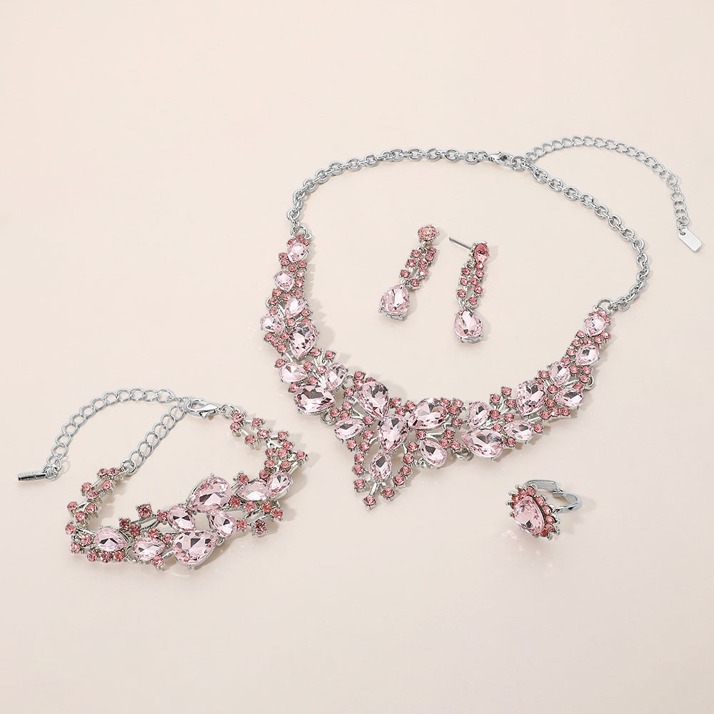 Pink crystal 4-piece jewelry set on silver with statement necklace, matching bracelet, drop earrings and adjustable ring arranged on a light beige background.