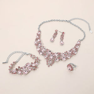 Pink crystal 4-piece jewelry set on silver with statement necklace, matching bracelet, drop earrings and adjustable ring arranged on a light beige background.