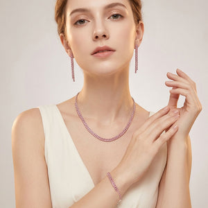Model wearing Pink crystal tennis-style jewelry set on silver-tone metal, featuring a single-row necklace, matching bracelet, and slender straight dangle earrings. Necklace measures 16 inches plus a 4-inch extender, bracelet 7 inches plus a 2-inch extender, and earrings 1.75 inches long. 