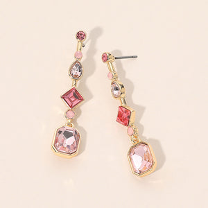 A pair of gold-plated dangle geometric earrings featuring pink and clear faceted gemstones in various shapes, displayed on a light beige background.