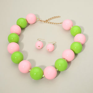 Pink and green chunky ball beaded statement necklace set with oversized round beads and matching earrings, displayed on a beige background.